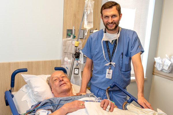 a man in a hospital bed and his doctor, a younger man wearing blue scrubs, standing next to him